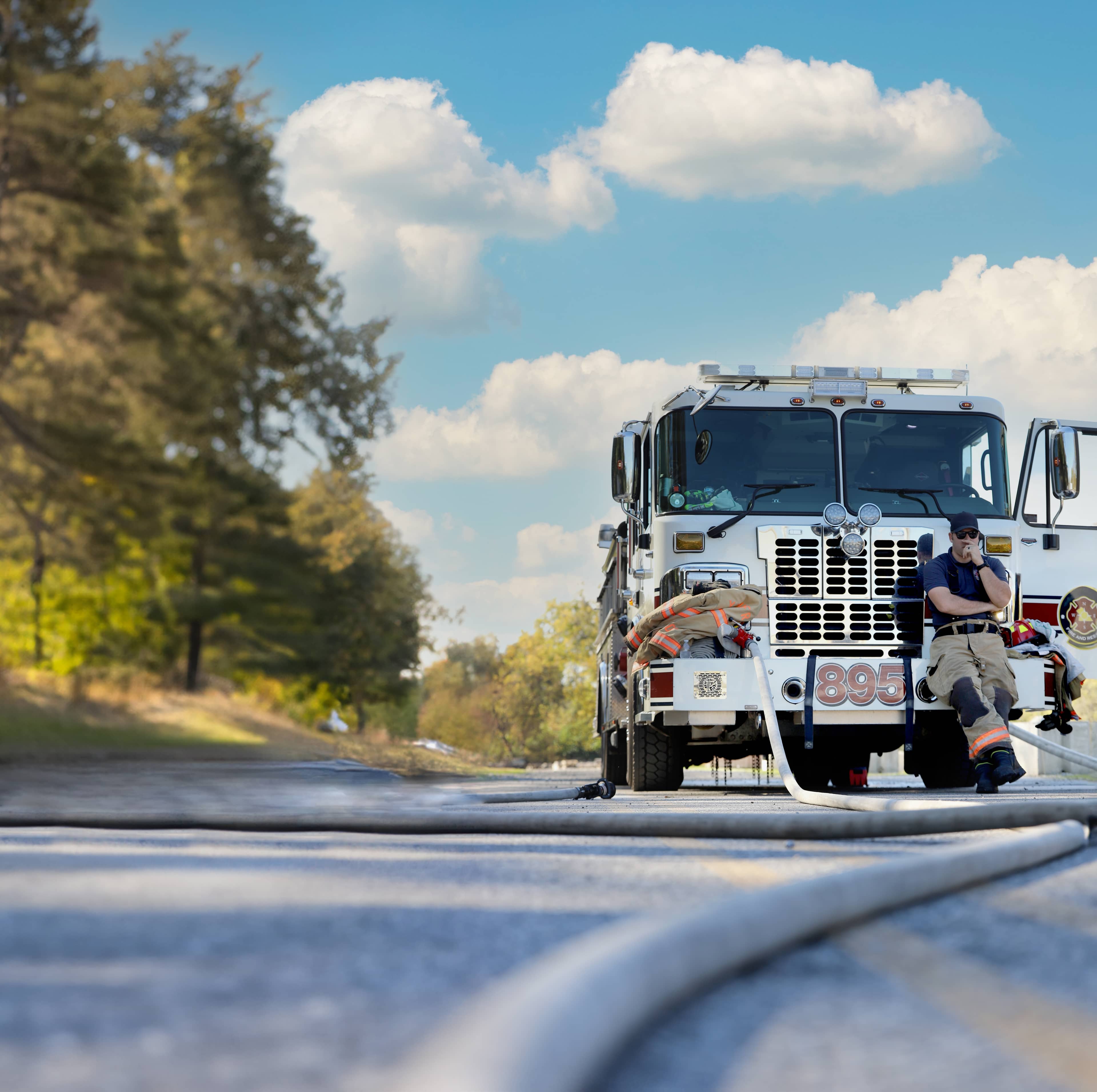 Firefighter in turnout gear resting against the front of a fire truck on a bright day