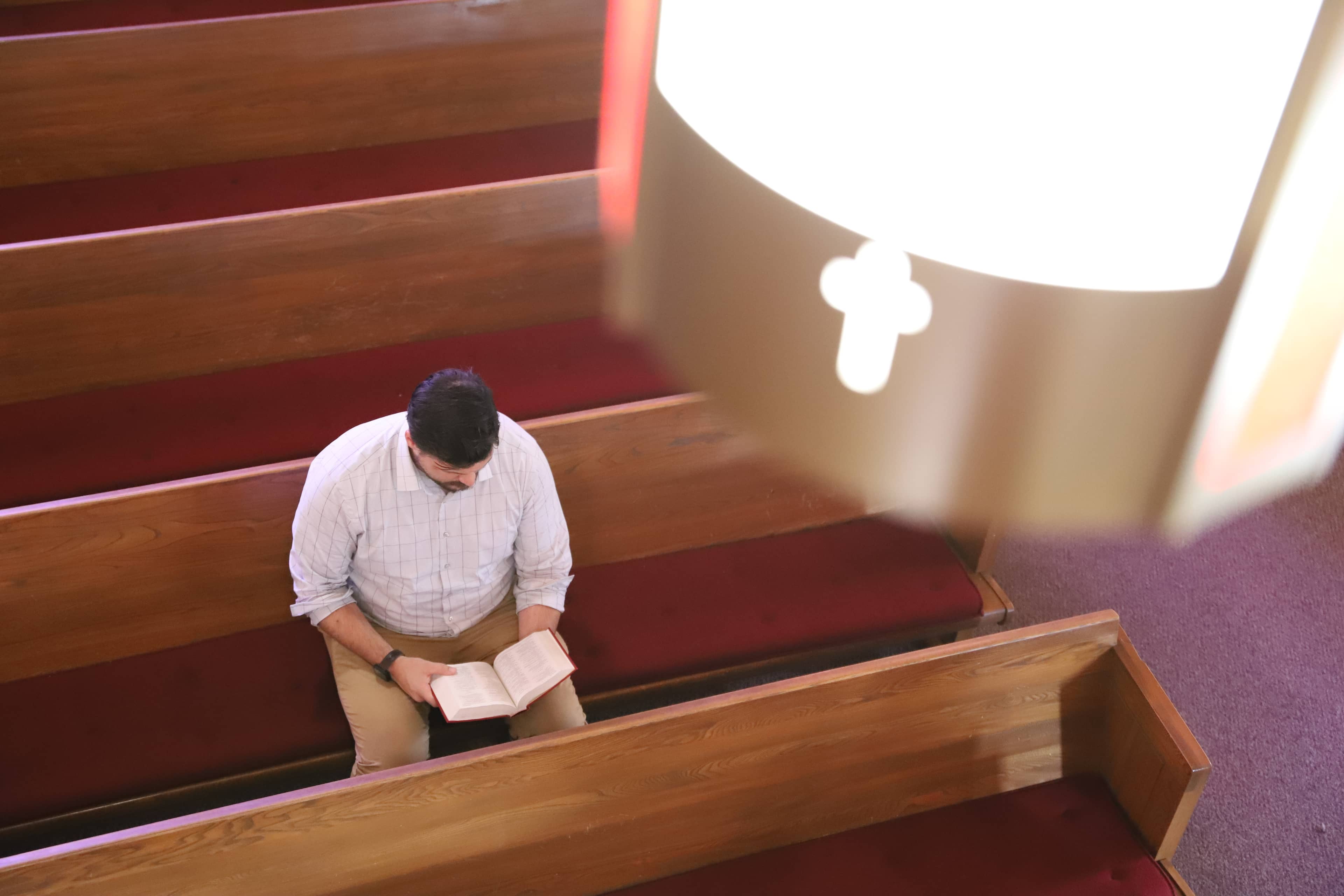 A man sitting alone in a church pew reading a Bible, viewed from above